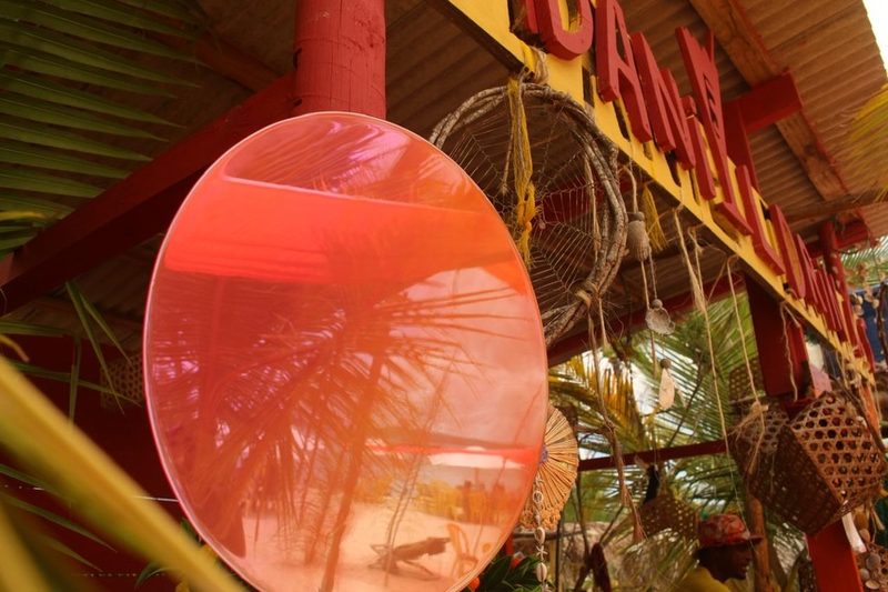 Espelho redondo rosa pendurado num quiosque de praia, refletindo coqueiros e a areia.